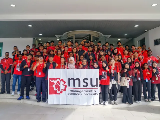 A large group of Management and Science University students in red shirts giving a thumbs-up on the steps of a PKT building behind an MSU banner