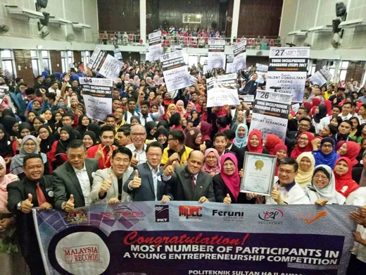 Hundreds of students and guests in a hall holding team placards behind a banner reading Most Number of Participants in a Young Entrepreneurship Competition, Malaysia Book of Records, with the PKT logo among the partners